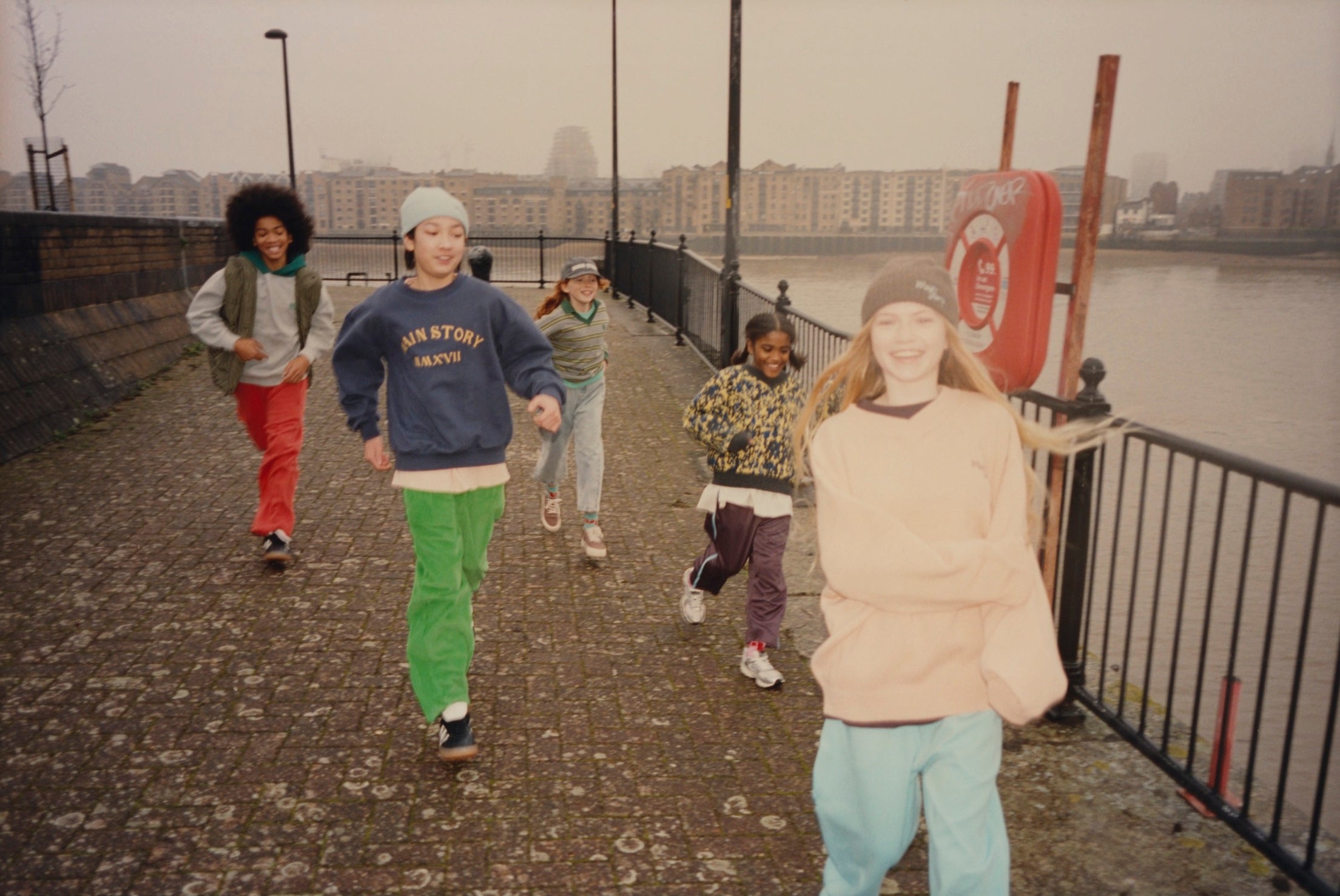 Five children running and playing on a paved riverside walkway with a black metal railing, water, and buildings in the background under an overcast sky.